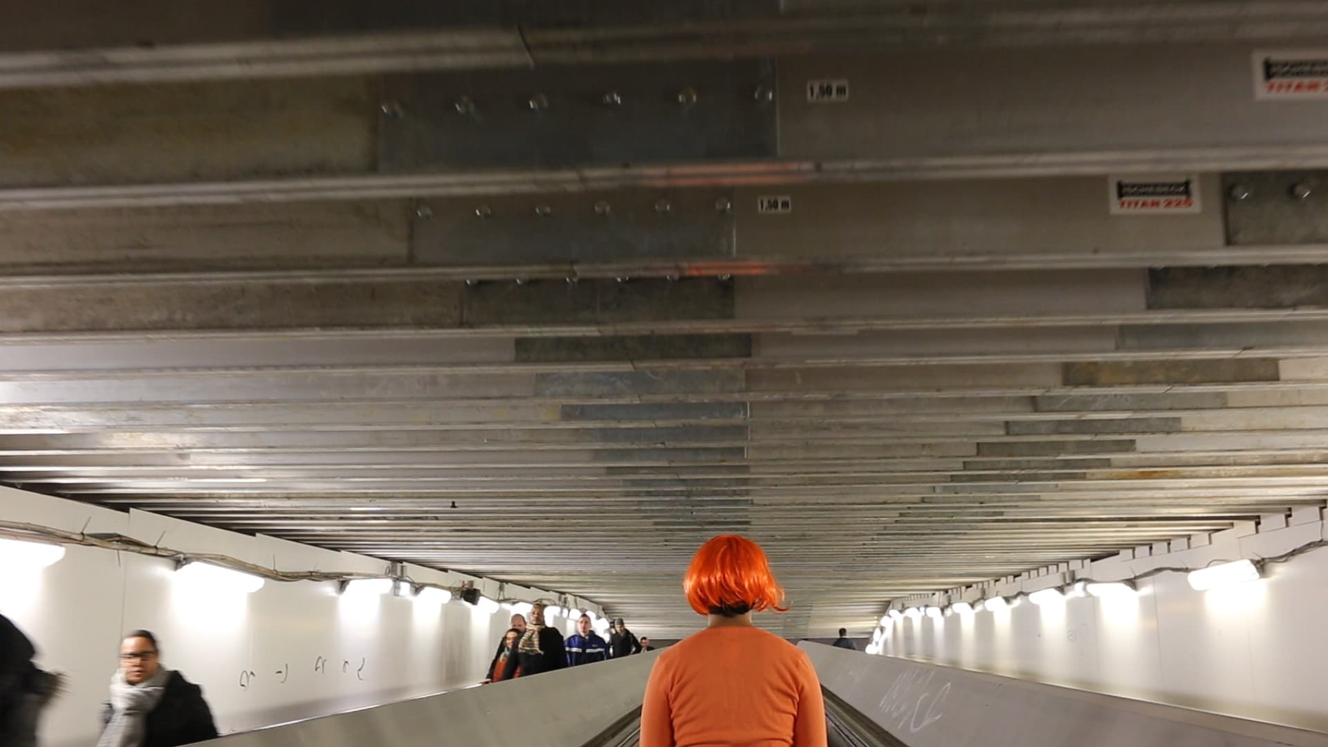 Still from the film Network/Intersect, by Ollie Palmer. The image depicts a person wearing an orange wig on an escalator.